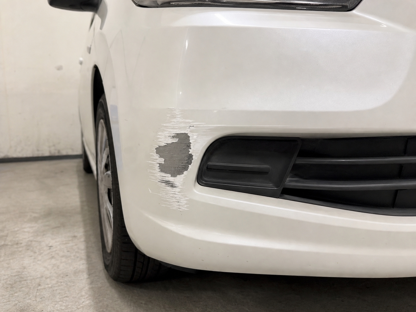 白いコンパクトカーのフロントバンパー左側。こぶし大の擦りキズと塗装剥がれがある修理前の状態